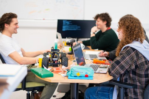 Three students round a desk smiling whilst working on a project