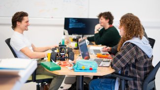 Three students round a desk smiling whilst working on a project
